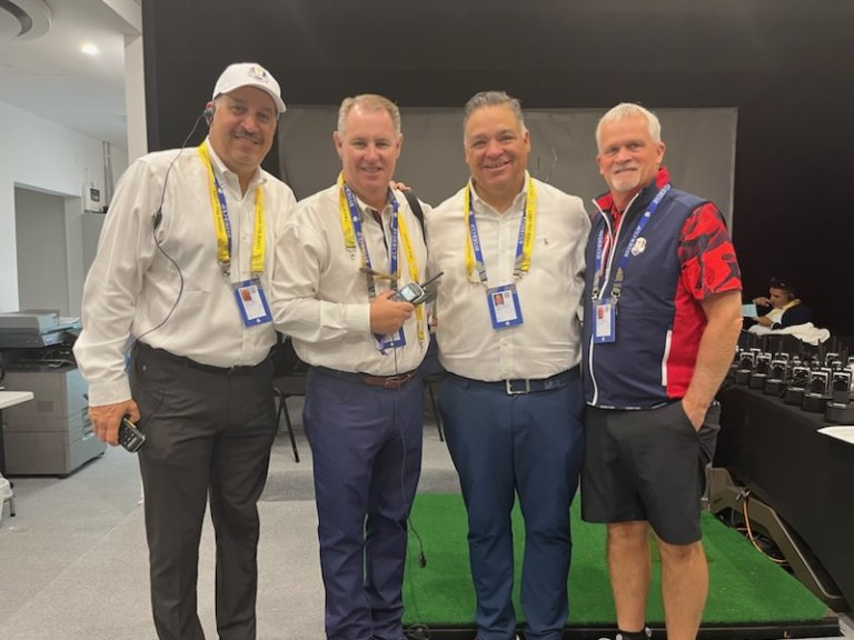 Four men are standing next to each other and smiling. The first three have beige jackets on and lanyards.