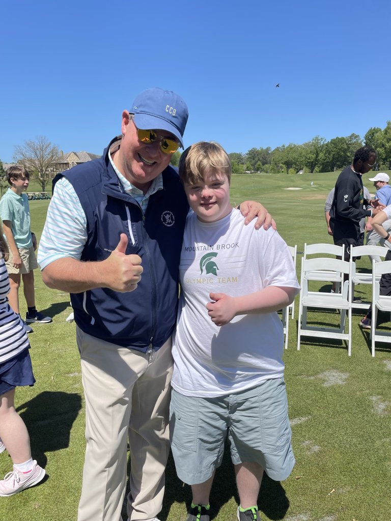 Eric hugs a boy with special needs on a golf course. They are both smiling and giving a thumbs up.