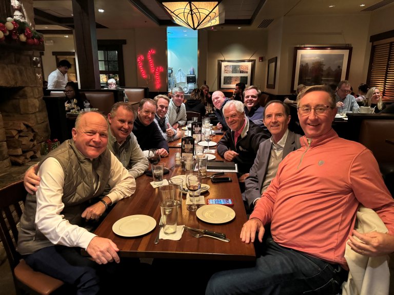 Ten men are sitting at a table after eating a meal. They are leaning in and smiling.